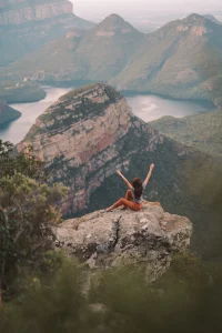 A Woman Raising Her Both Arms while sitting on a top of rock at mountain