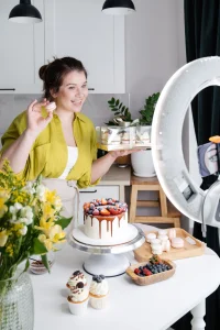 Smiling woman showing assorted yummy homemade cakes while recording vlog by iPhone set with tripod