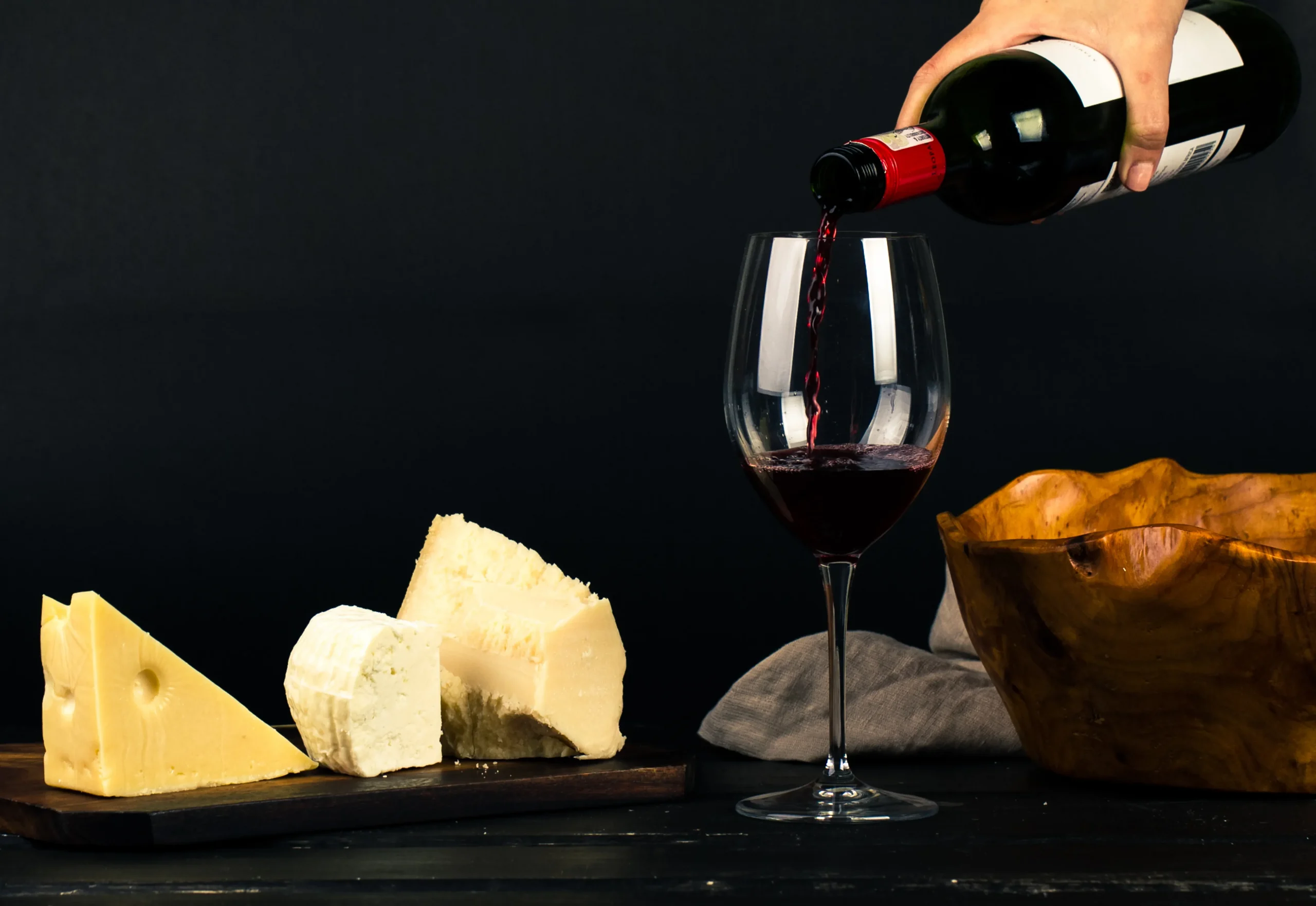 A Chiaroscuro photo of person pouring wine into glass besides some cheese pairings

