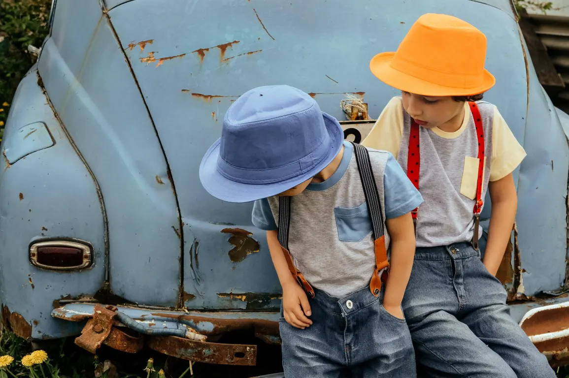 Two boys wear in blue hat and orange hats and sitting on a blue car bumper